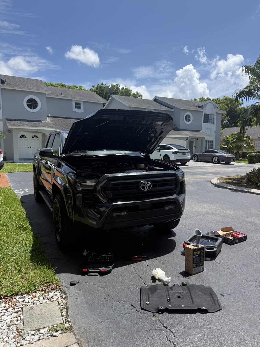 Tacoma with hood and skid plate off during oil change, tools and oil laid out on the driveway
